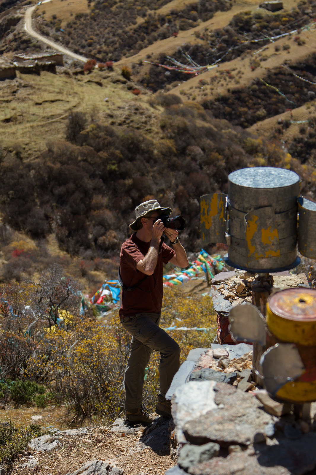 Man in a sun hat photographing with a DSLR on a Tibetan hillside strung with prayer flags.