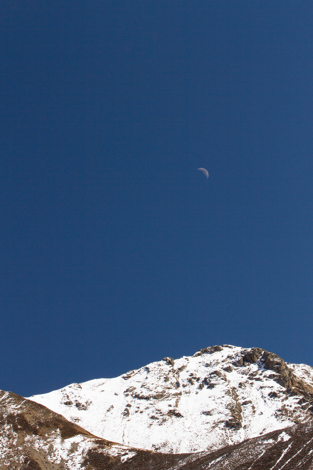A half moon in a deep blue sky above a snow-dusted mountain ridge.