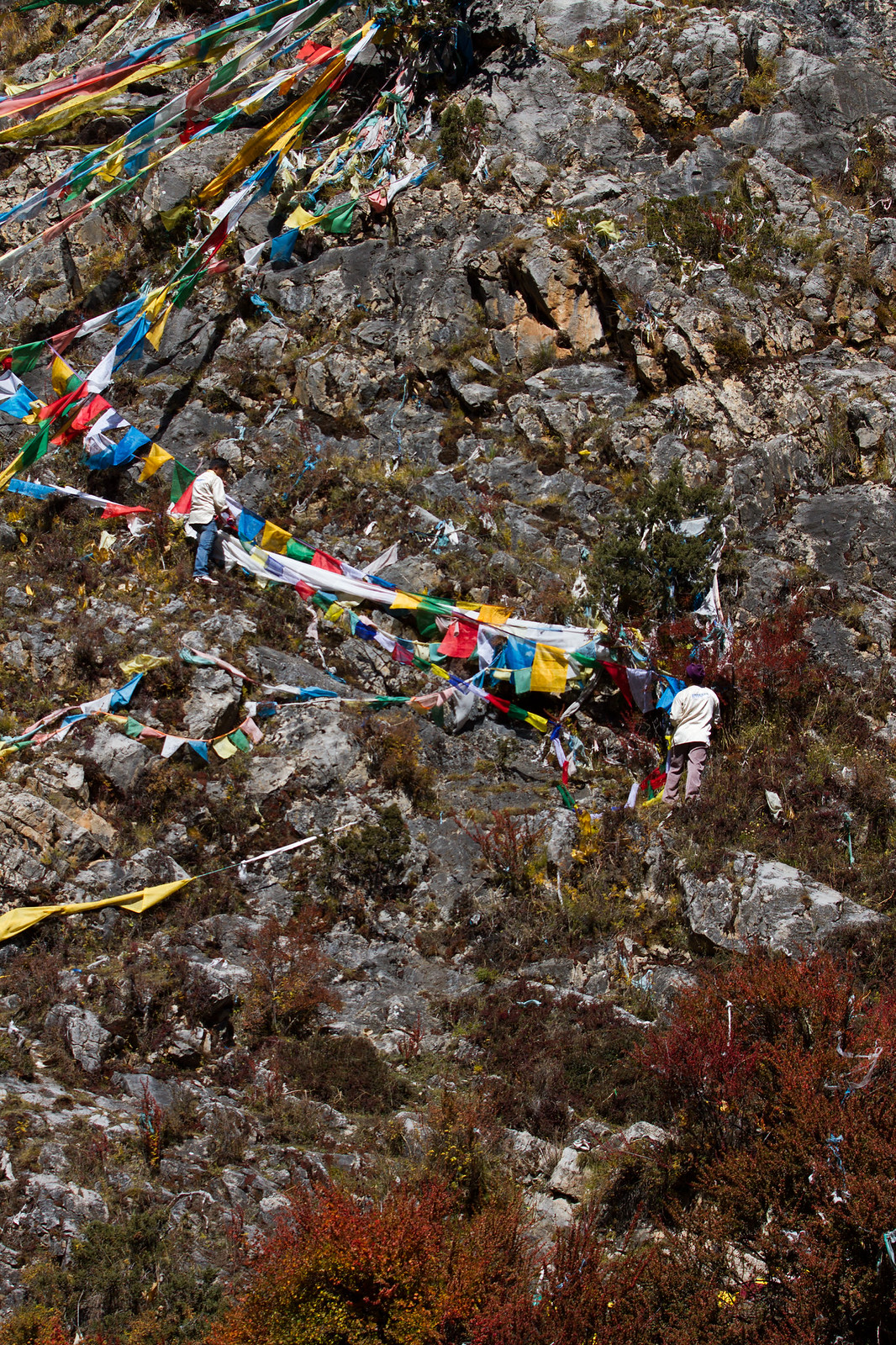 Two people string colorful Tibetan prayer flags across a rugged rocky mountainside.
