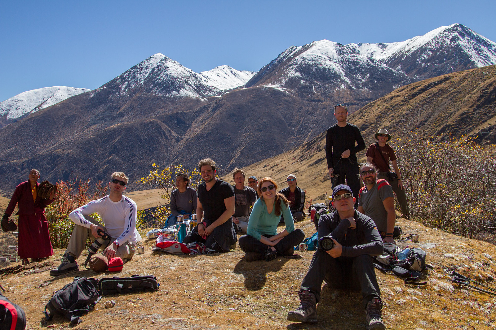 A group of trekkers and a red-robed monk pause on a hillside below snow-capped Himalayan peaks.