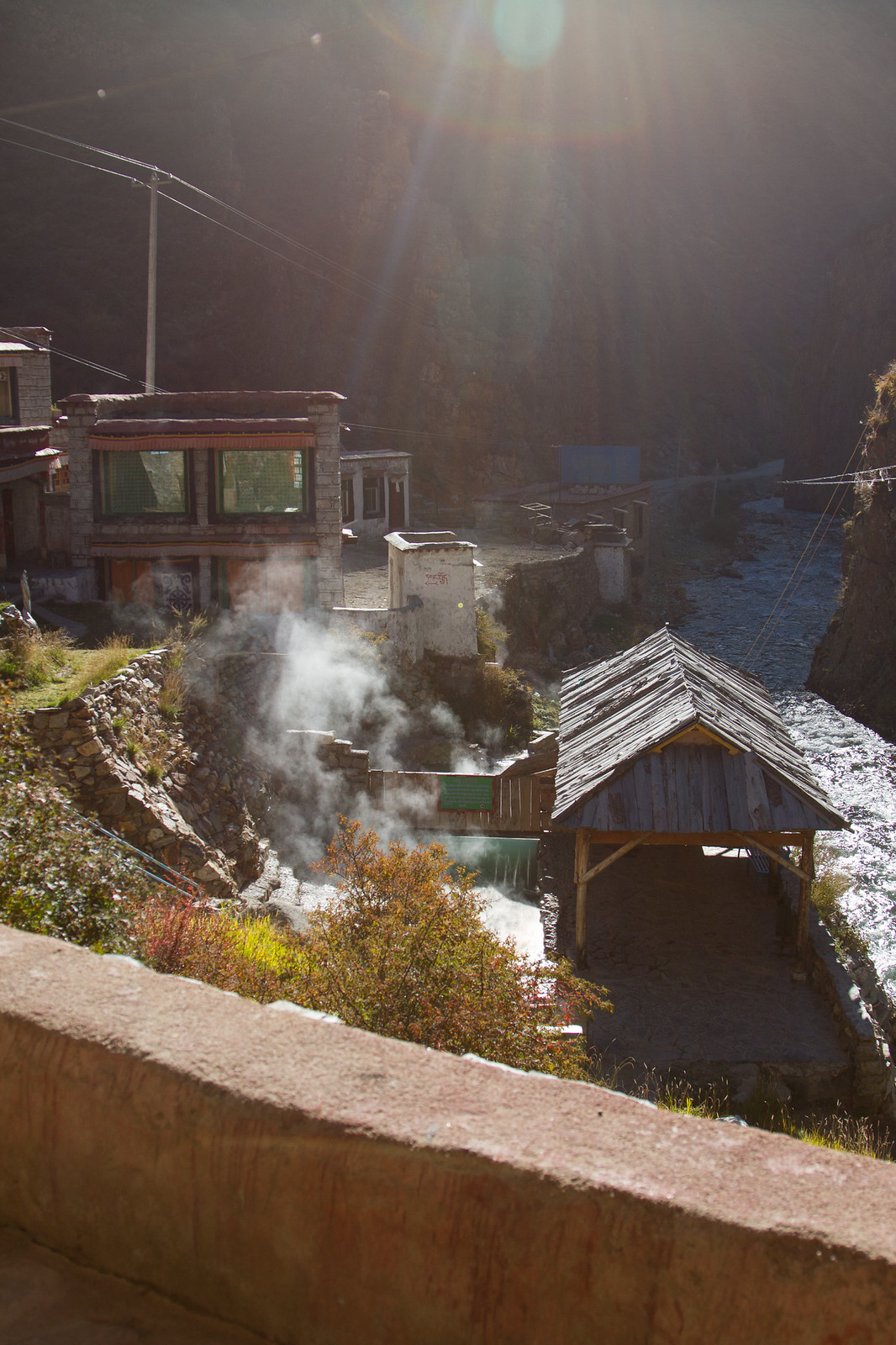 Steam rises from a hot spring beside a river and Tibetan stone buildings, backlit by low sun.