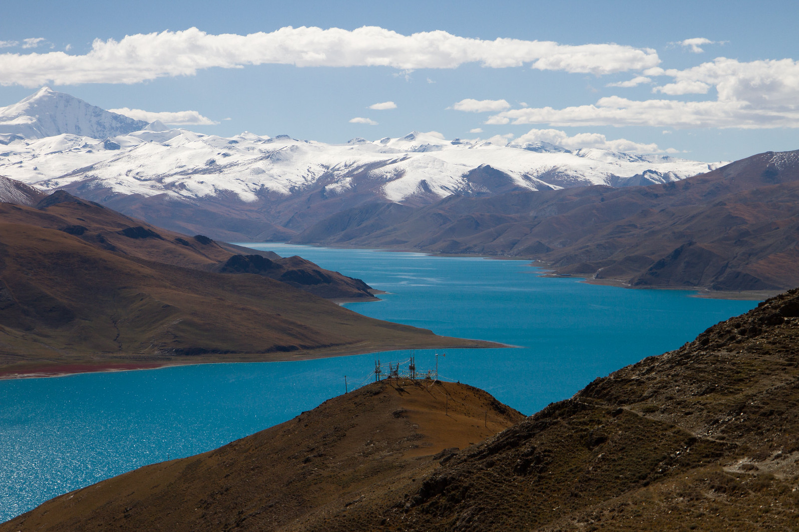 Turquoise Yamdrok Lake winding between barren brown hills below snow-capped Himalayan peaks in Tibet.