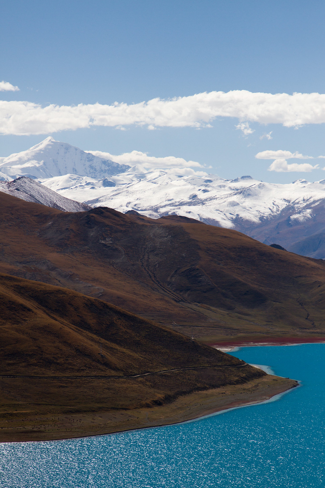 Turquoise waters of Yamdrok Lake below brown Tibetan hills and snow-capped Himalayan peaks under a blue sky.