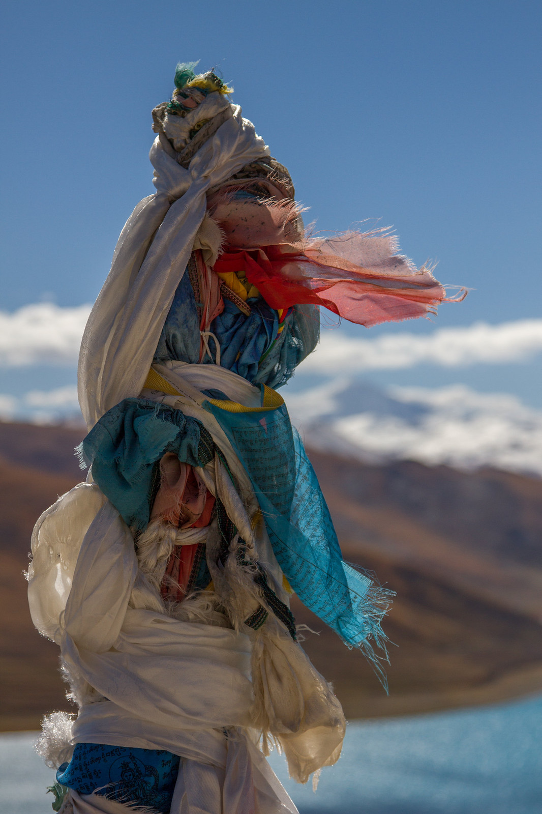Weathered Tibetan prayer flags and white khata scarves flutter in the wind above a lake and snow-capped peak.