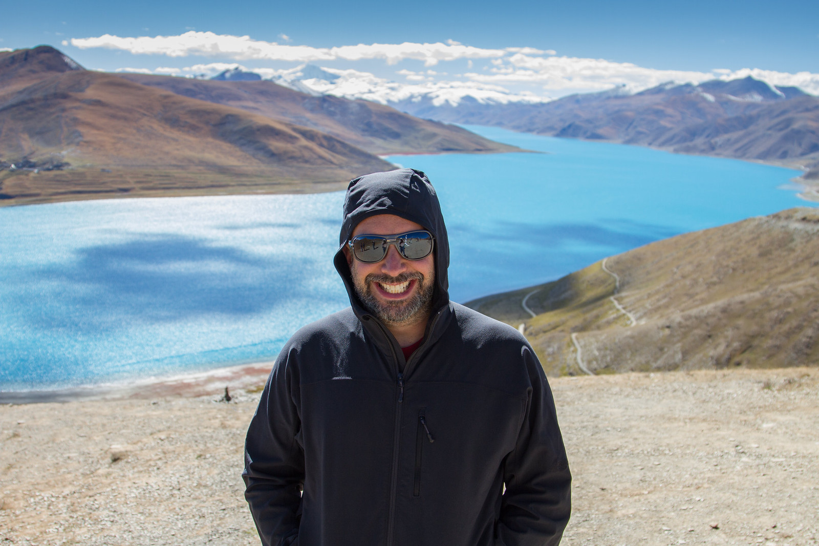 A smiling bearded man in a black hooded jacket and sunglasses stands above the turquoise Yamdrok Lake in Tibet.