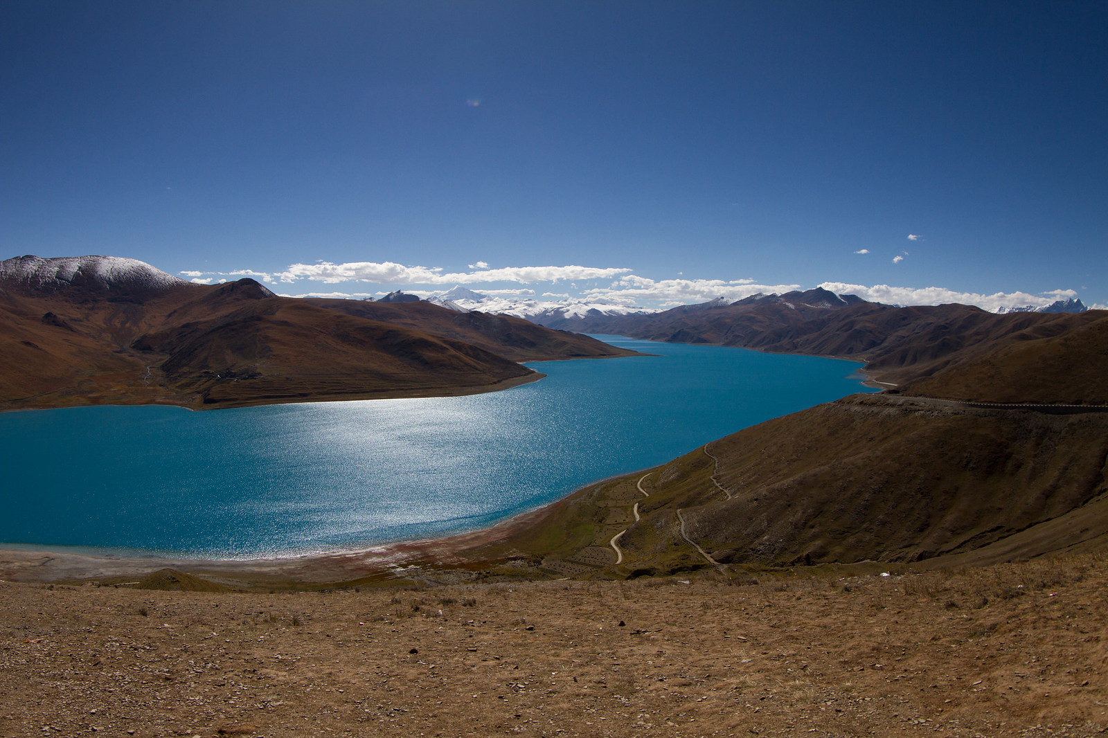 Turquoise waters of Yamdrok Lake winding between barren brown hills toward snowcapped Himalayan peaks under a deep blue sky.