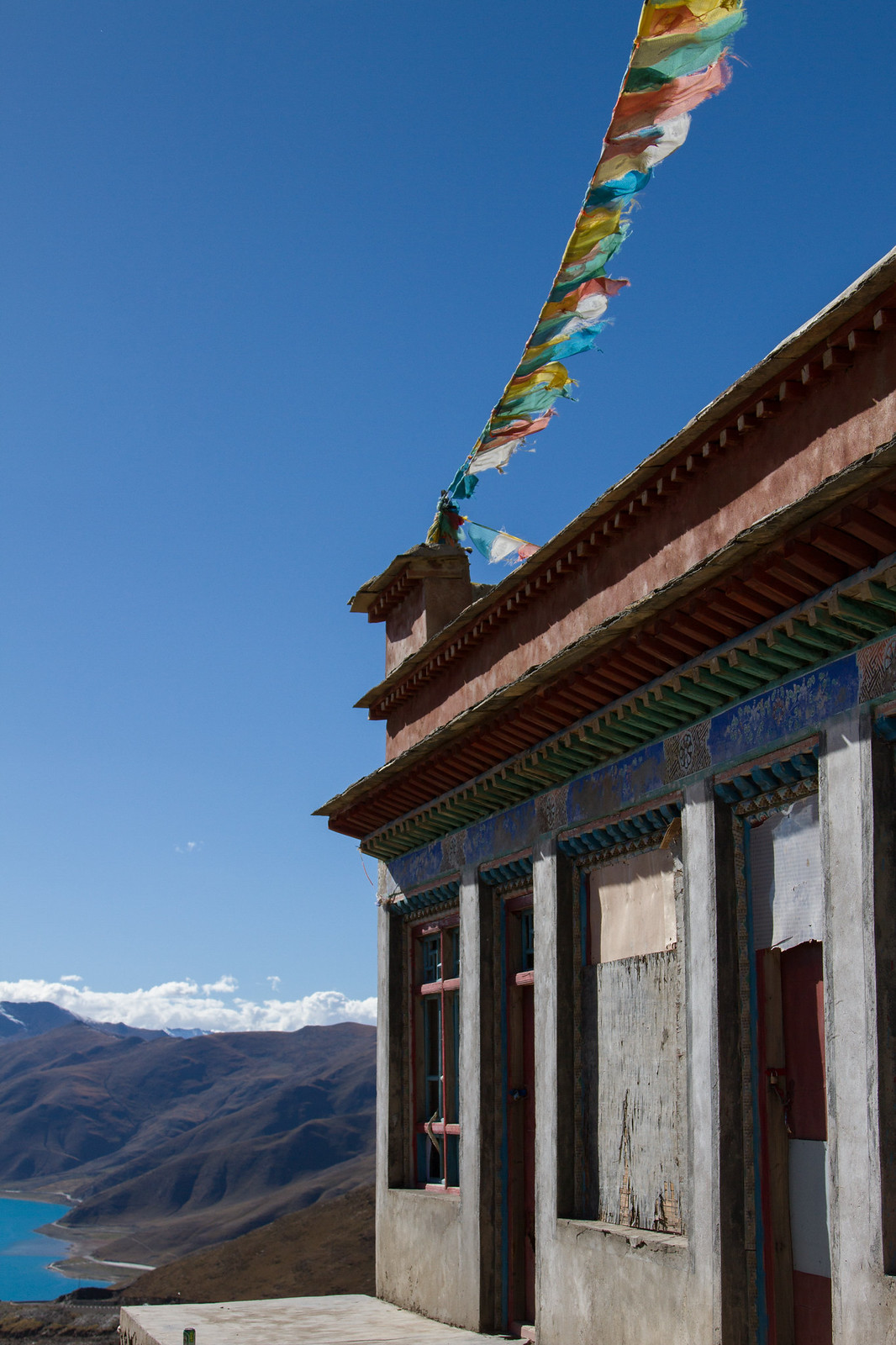 Colorful Tibetan prayer flags strung from a monastery rooftop above a turquoise lake and mountains.