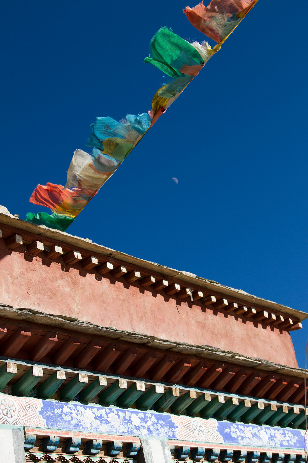 Colorful Tibetan prayer flags strung above the ornately painted eaves of a red monastery against a deep blue sky.