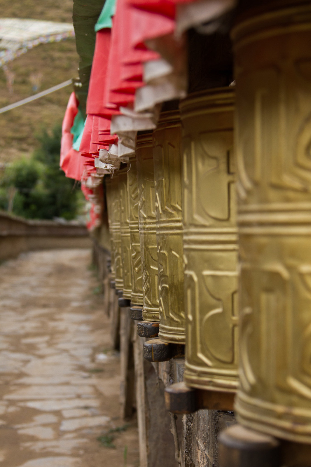 A row of golden Tibetan prayer wheels topped with red and green cloth along a stone path.