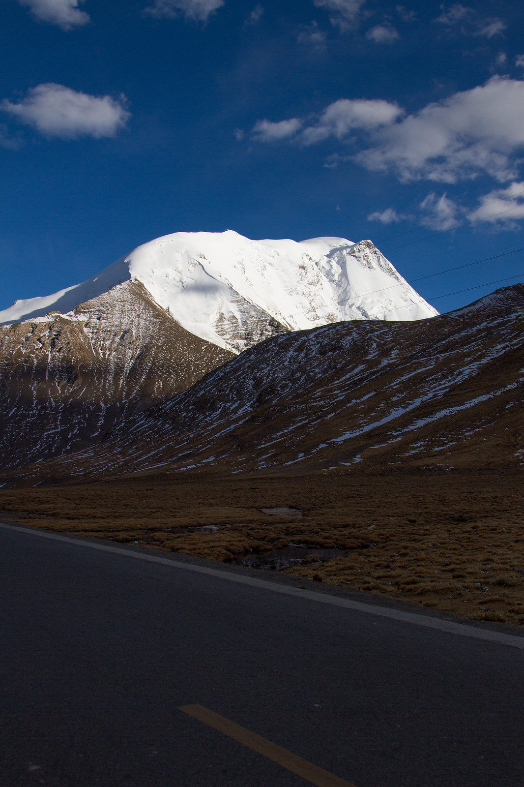 Empty highway curves past a snow-capped Himalayan peak under a deep blue sky in Tibet.