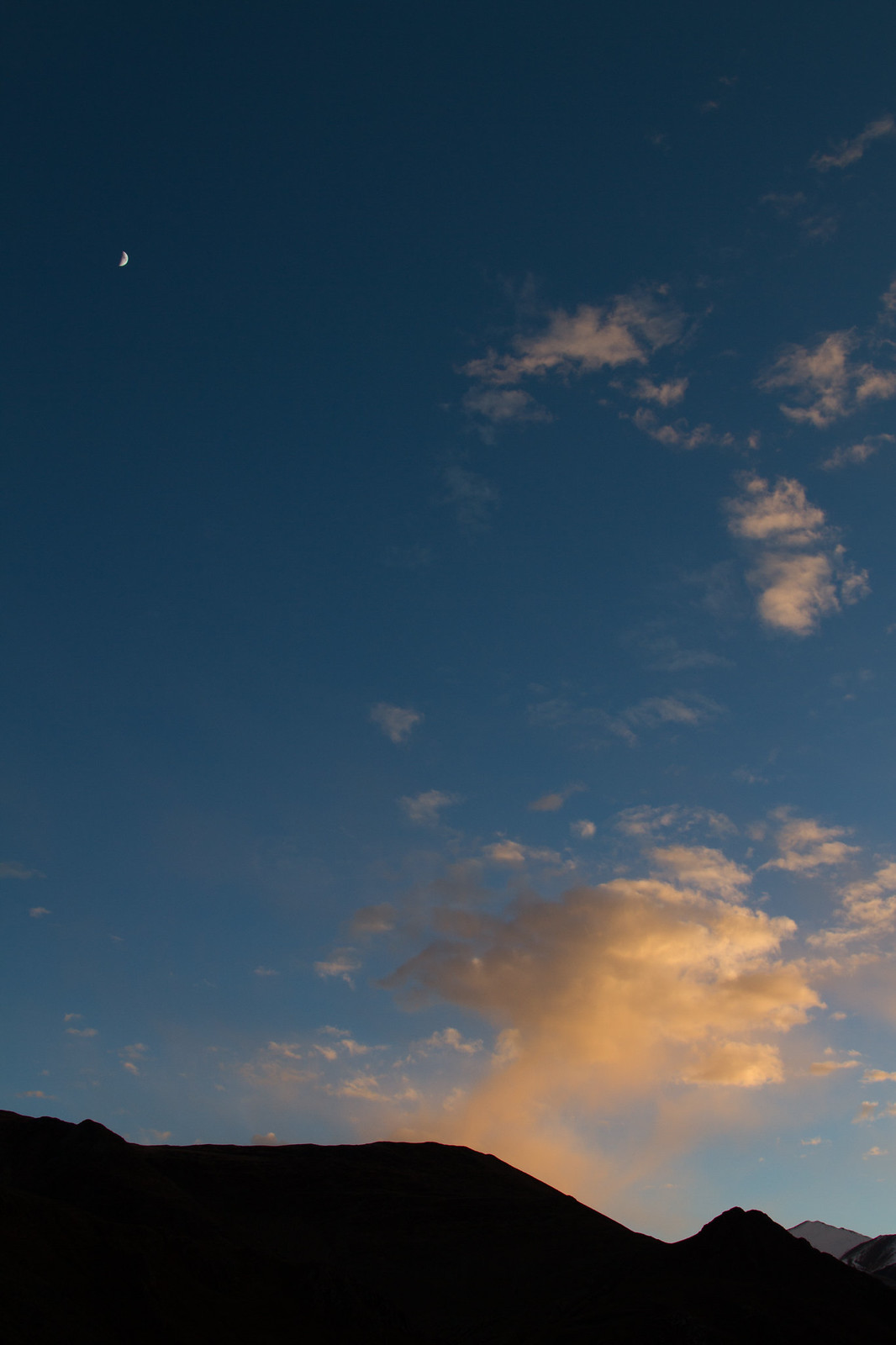 A crescent moon and golden sunset-lit clouds over silhouetted mountain ridges in a deep blue sky.