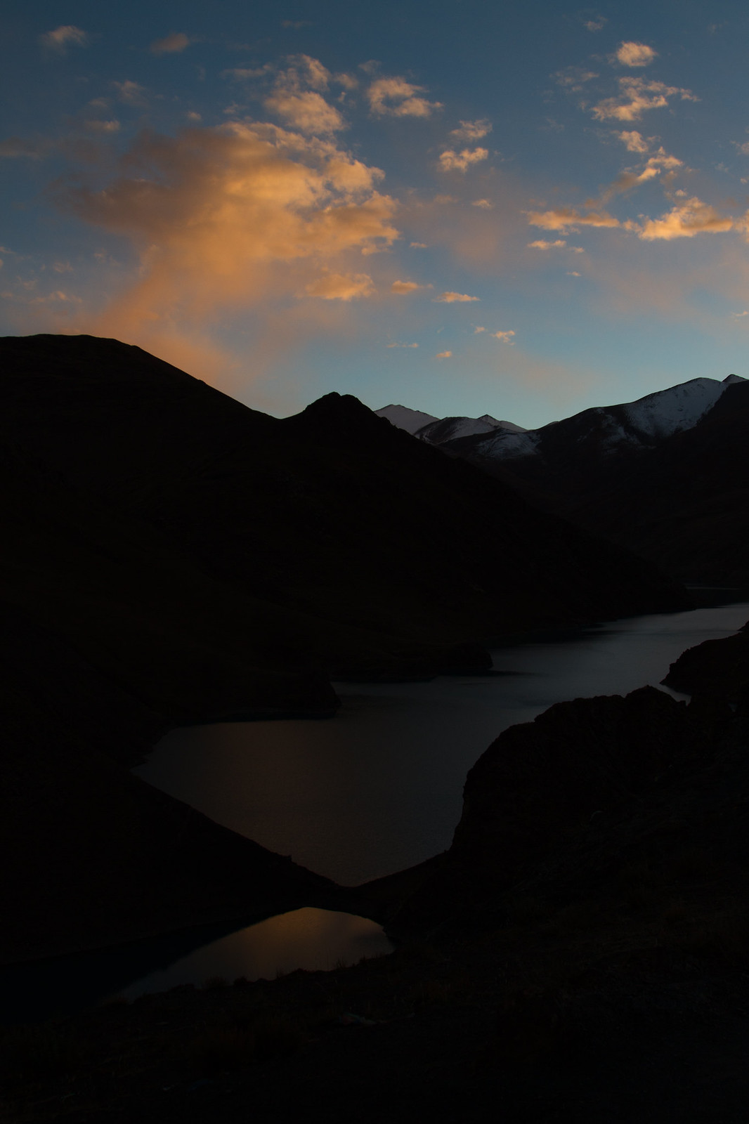 Silhouetted mountains and a still lake at dusk, with orange-lit clouds glowing over distant snow-capped peaks.