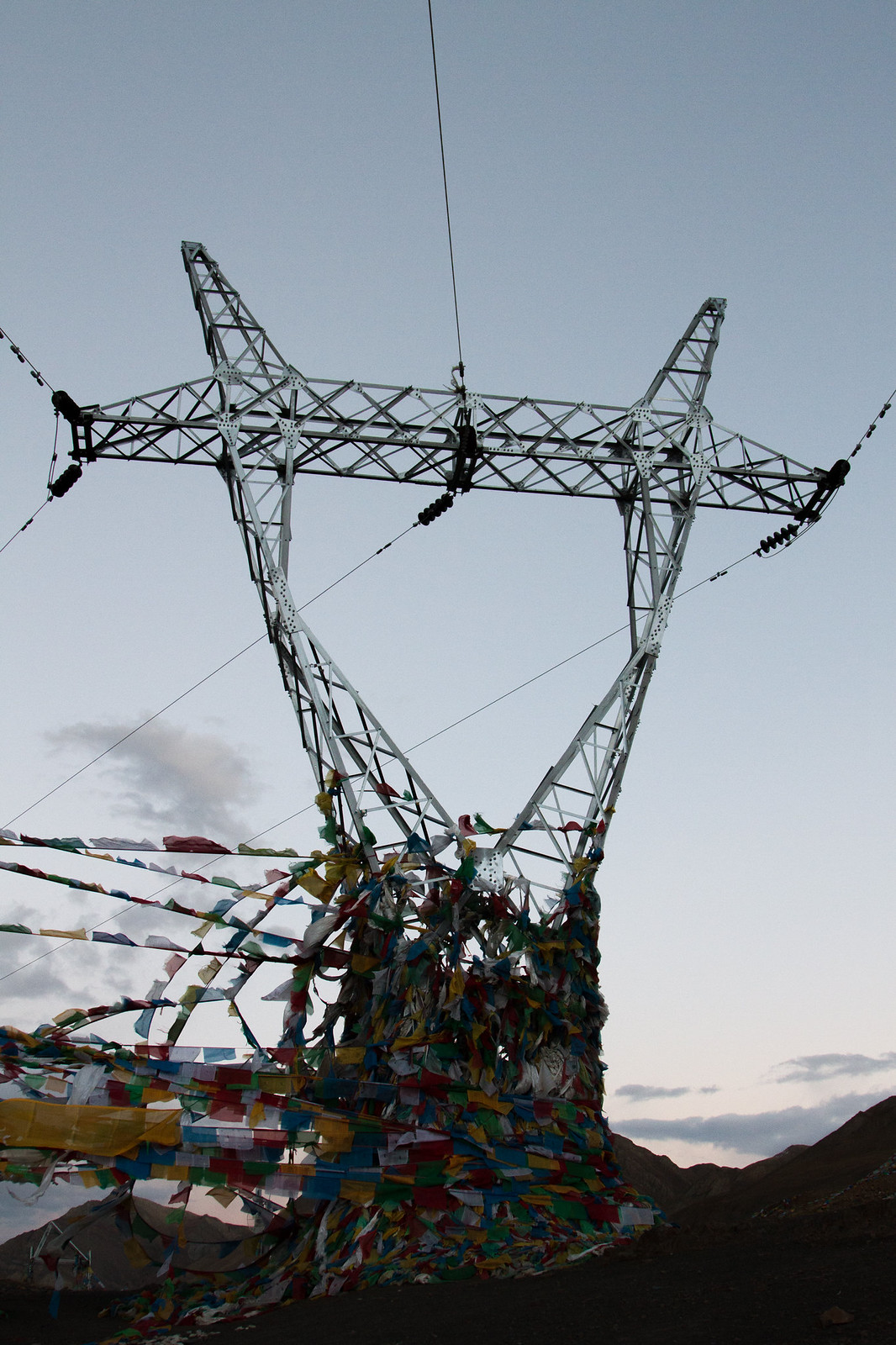 Colorful Tibetan prayer flags draped around the base of an electricity pylon at dusk in the mountains.