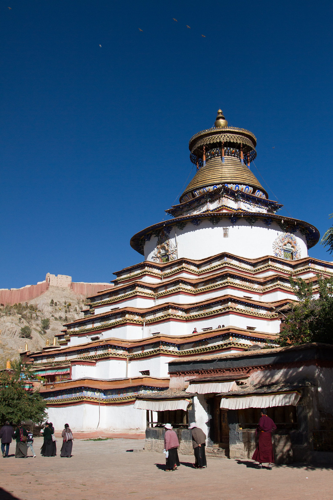 The tiered white-and-gold Kumbum stupa at Palcho Monastery with pilgrims walking below against a deep blue sky.
