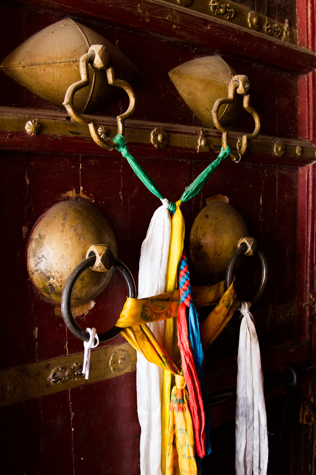 Ornate brass door knockers on a red Tibetan temple door draped with white, yellow, and colorful prayer scarves.