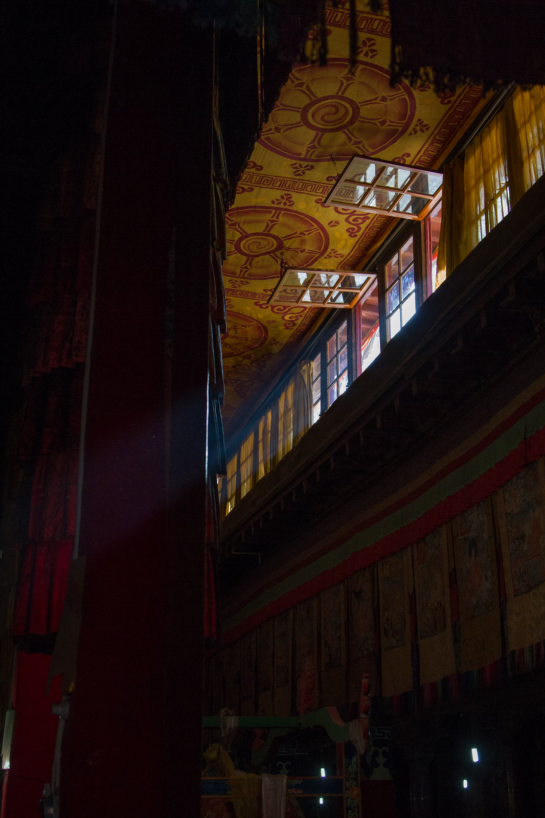 Light streams through open windows onto a Tibetan monastery's ornate painted ceiling and richly decorated interior walls.