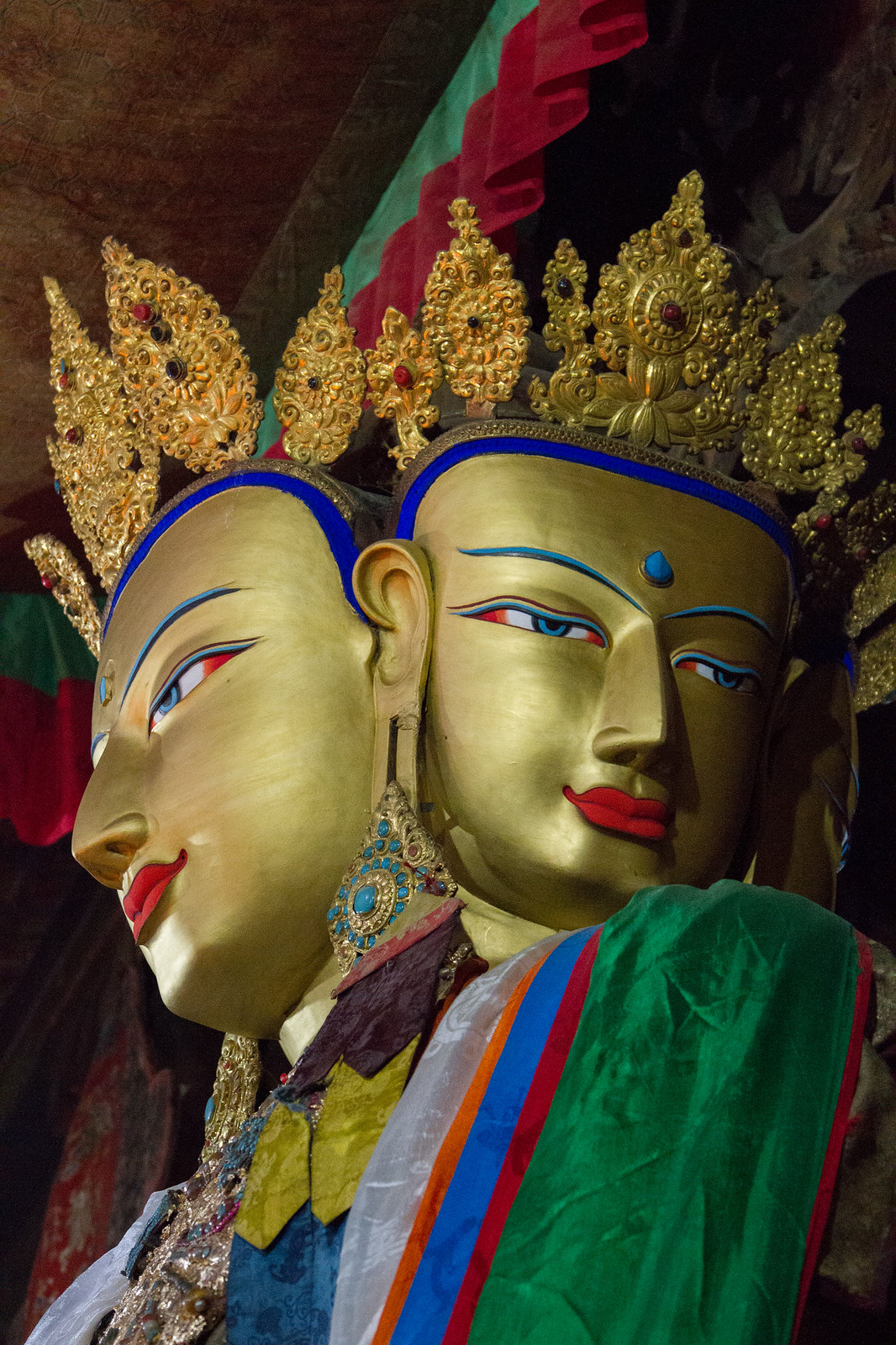 Gilded multi-faced Buddha statue with jeweled gold crowns, blue-lined eyes, and colorful silk robes in a Tibetan temple