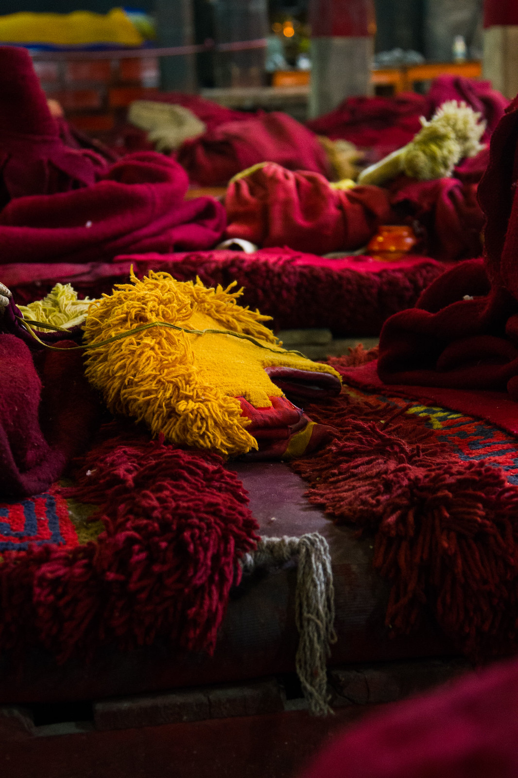 Crimson monks' robes and yellow crested hats laid out on cushioned benches inside a dim Tibetan monastery hall.
