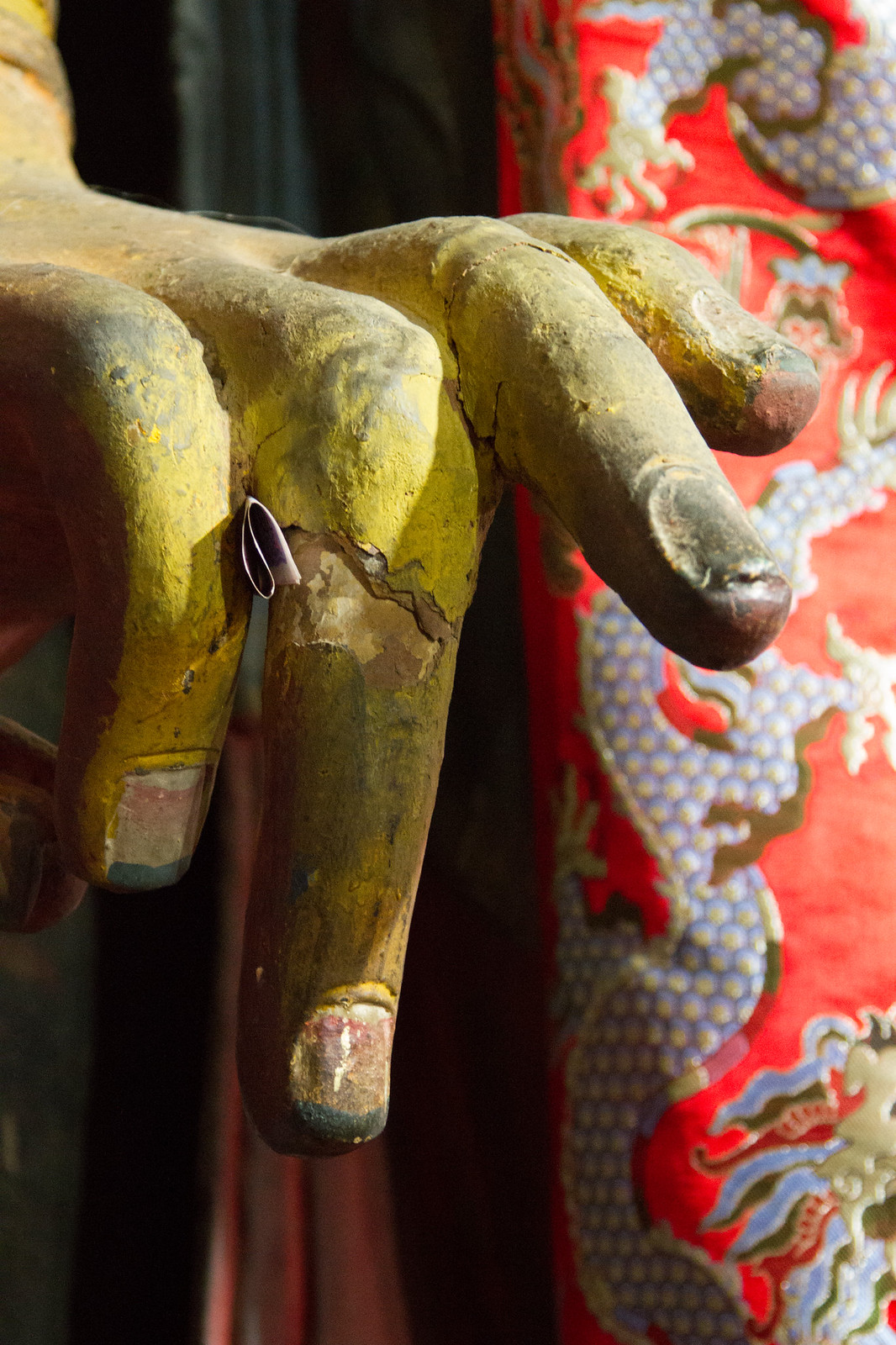 Close-up of a weathered gilded Buddha statue's hand beside red silk brocade with dragon motifs.