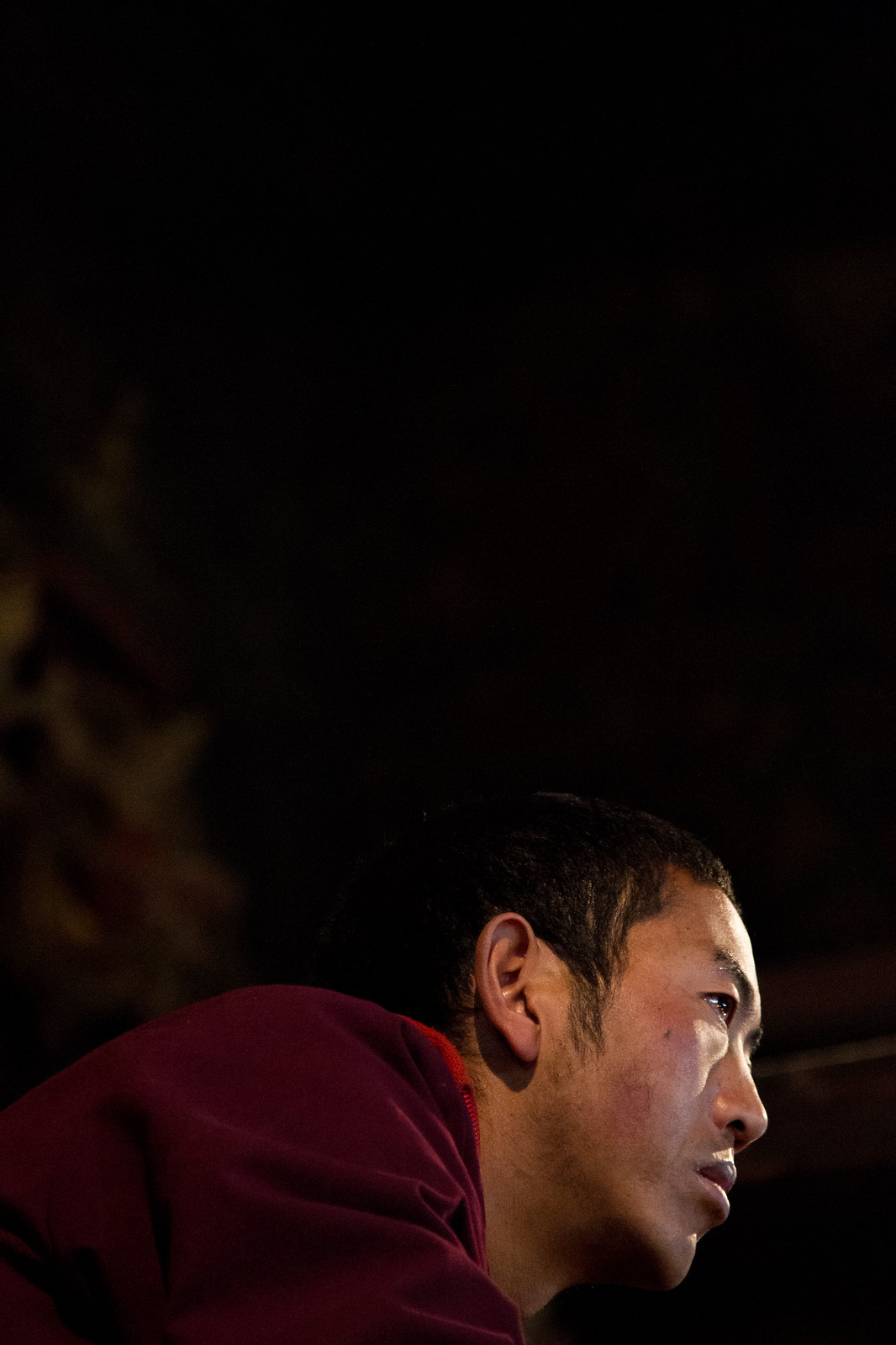 A monk in maroon robes gazes intently to the side, lit softly against deep shadow.