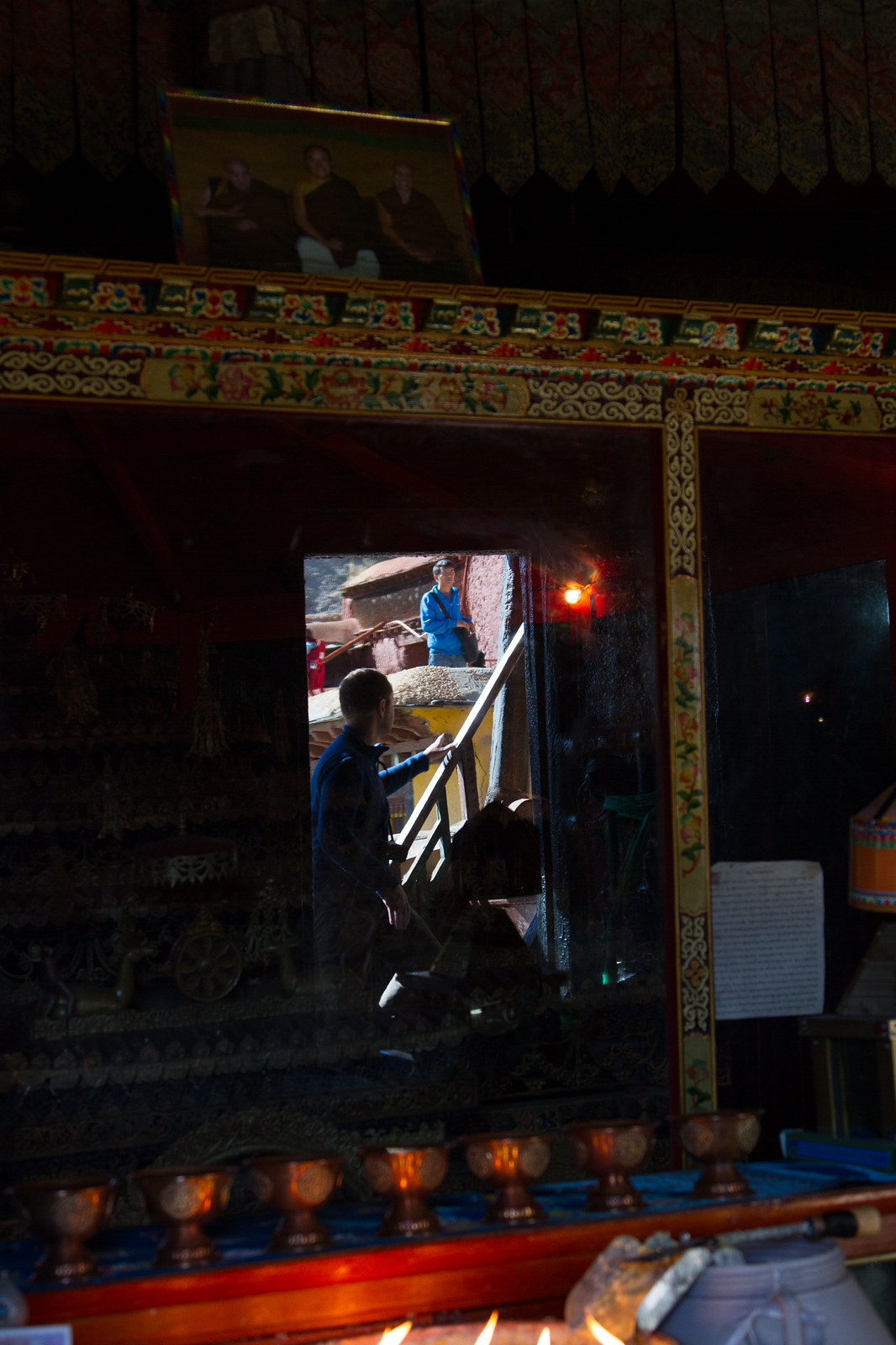 Reflections of visitors and a sunlit courtyard in the glass of a dim, ornately painted Tibetan monastery shrine.