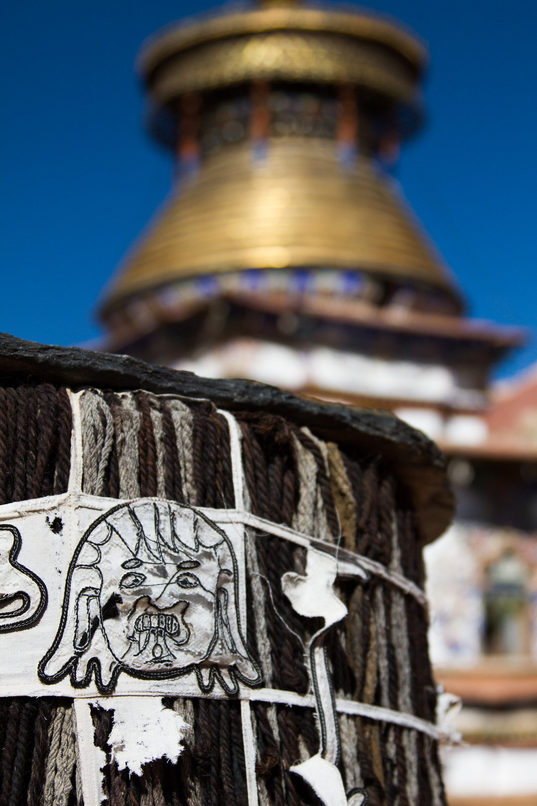 A woven black yak-hair post with an embroidered face motif below a golden Tibetan monastery roof against blue sky.