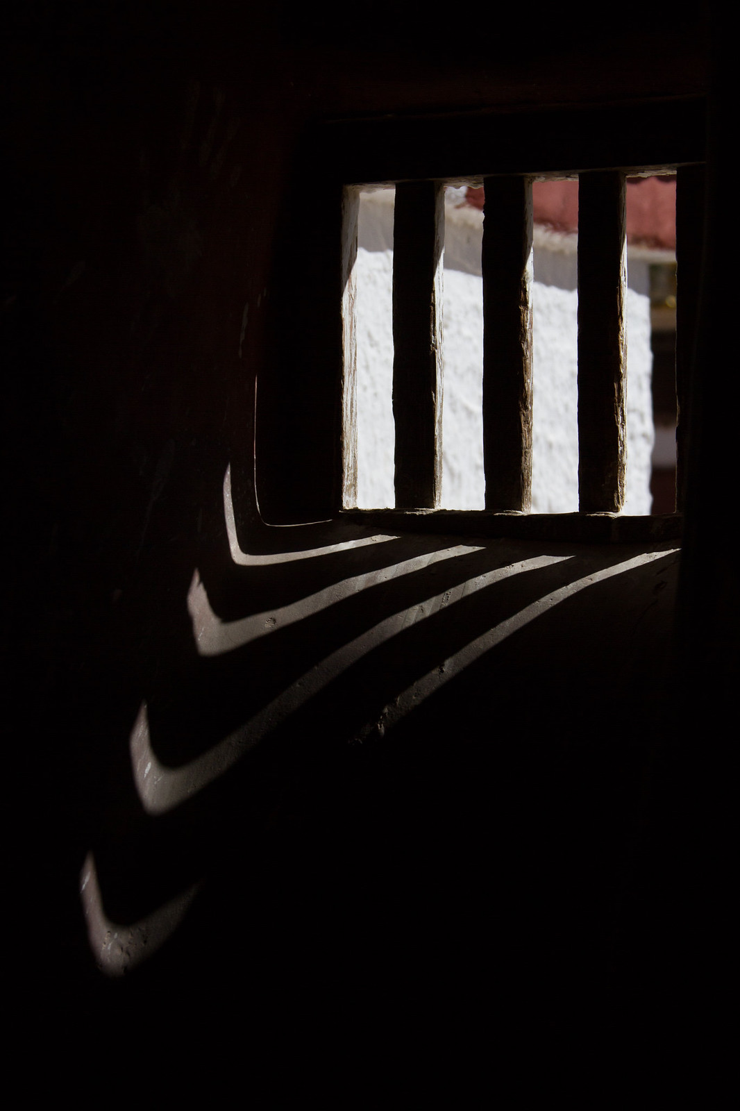 Sunlight streams through a barred window in a dark Tibetan monastery wall, casting striped shadows on the sill.