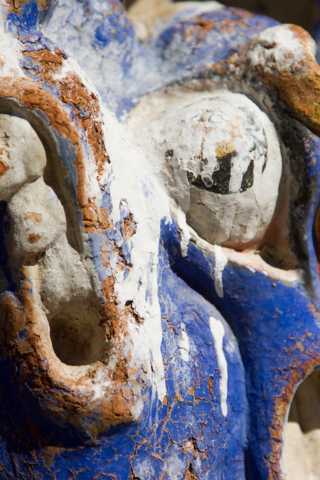 Weathered blue, white, and terracotta painted stone statue with cracked, peeling surface in bright sunlight.