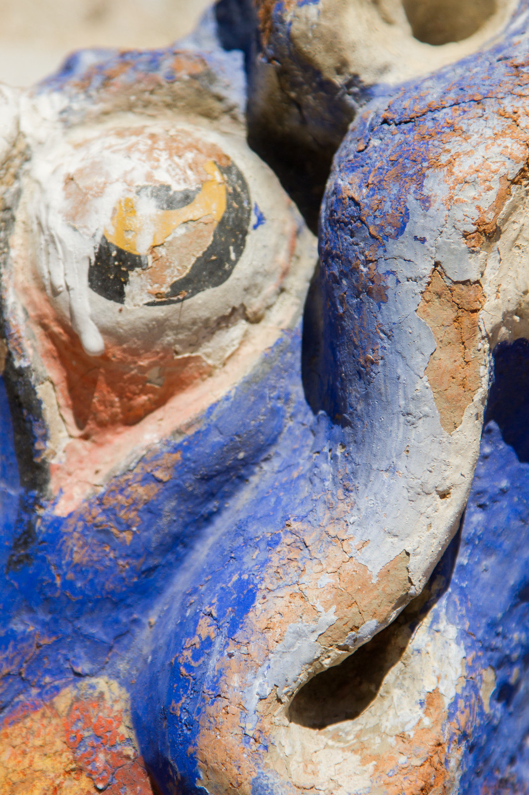 Close-up of a weathered blue and orange statue with a painted, cracked eye and peeling paint.
