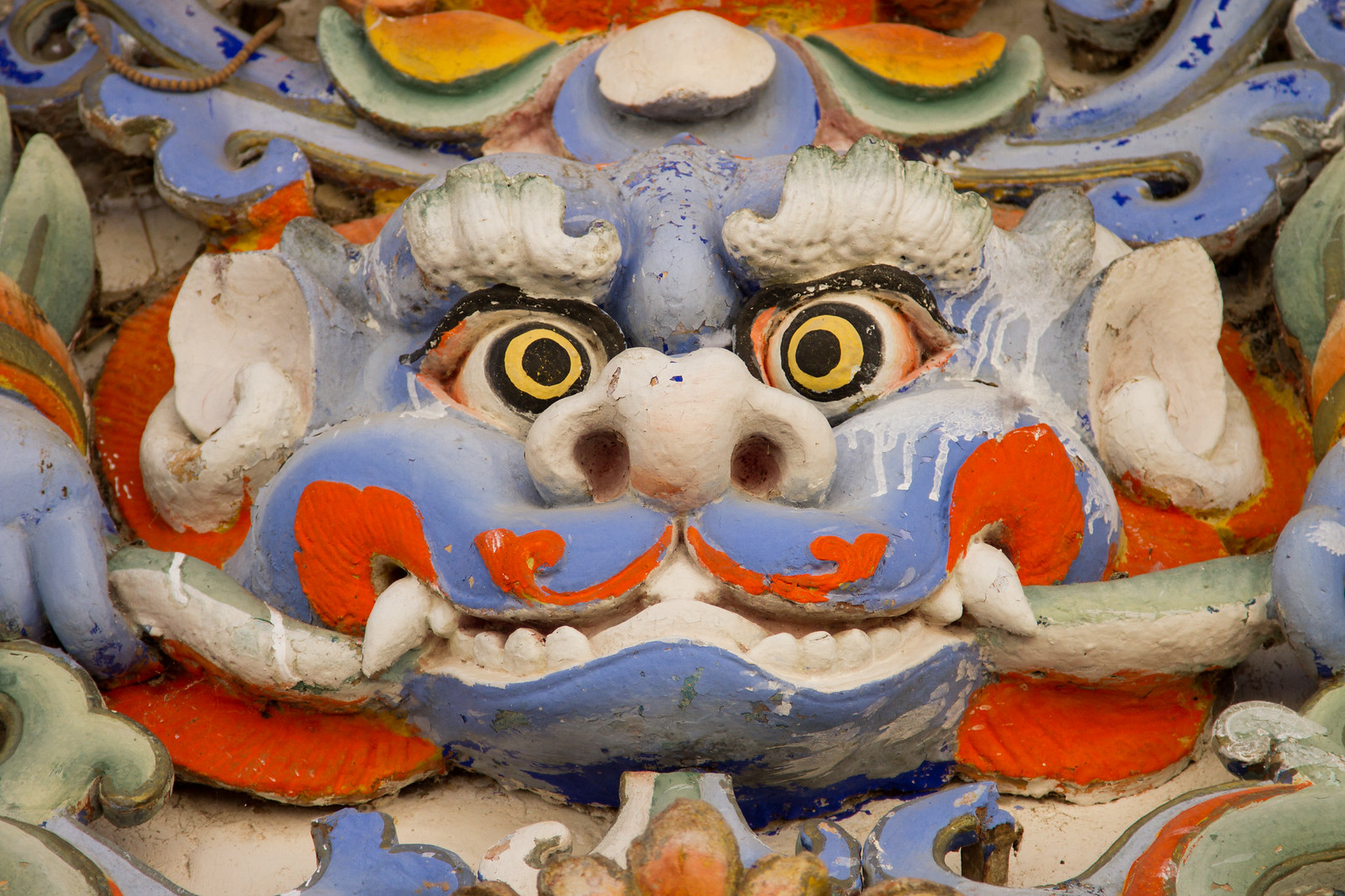 Ornate painted relief carving of a fierce blue Tibetan lion or dragon face with yellow eyes and orange flourishes.