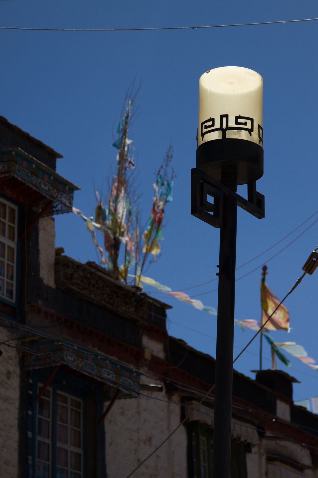 Ornate street lamp against blue sky, with a Tibetan building and colorful prayer flags behind it.