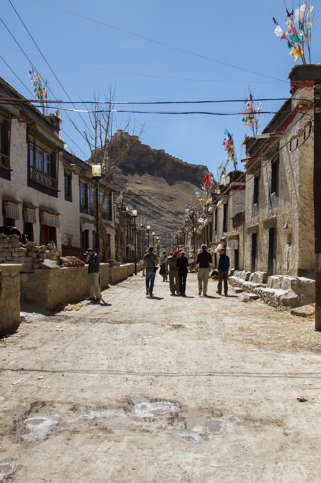 Tourists walk a dusty Tibetan village street lined with whitewashed prayer-flag-draped houses, a hilltop fortress ruin ahead.
