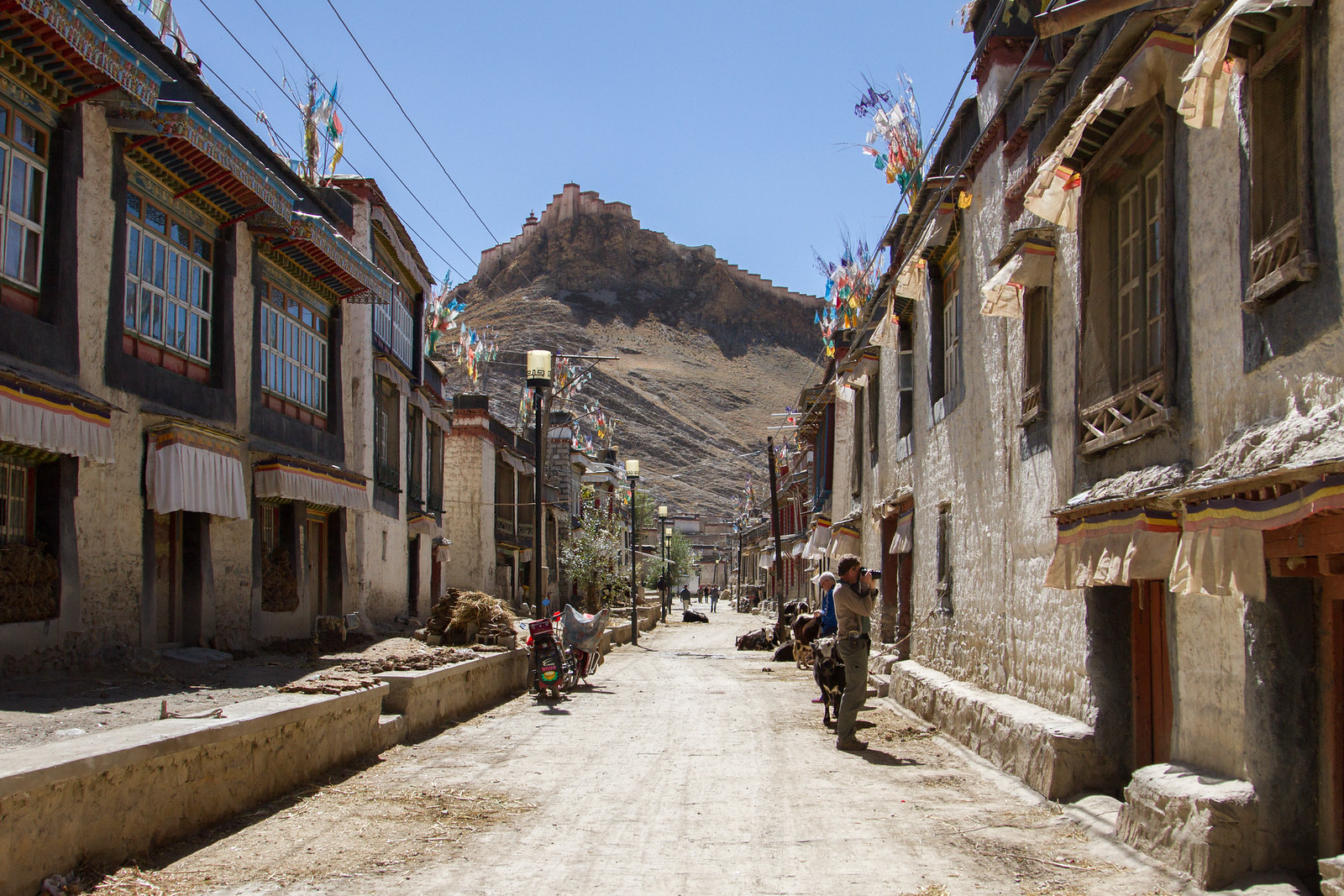 A dusty village street in Gyantse, Tibet, framed by whitewashed buildings and prayer flags, with a hilltop fort beyond.