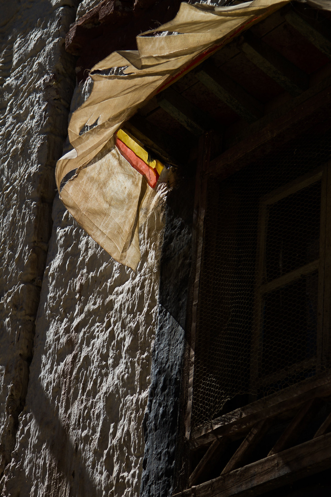 Weathered cloth awning over a wire-screened window on a sunlit whitewashed Tibetan monastery wall.