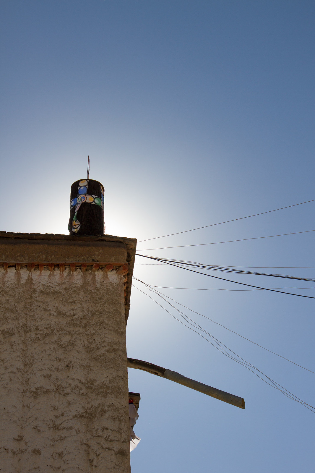 Painted cylindrical ornament atop a whitewashed Tibetan rooftop, backlit by sun with wires fanning across a clear blue sky.