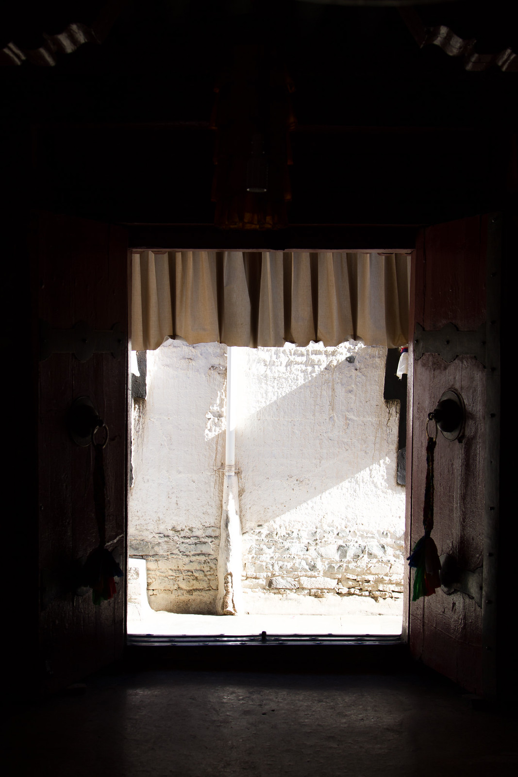View through a dark Tibetan monastery doorway to a sunlit whitewashed stone wall beyond.