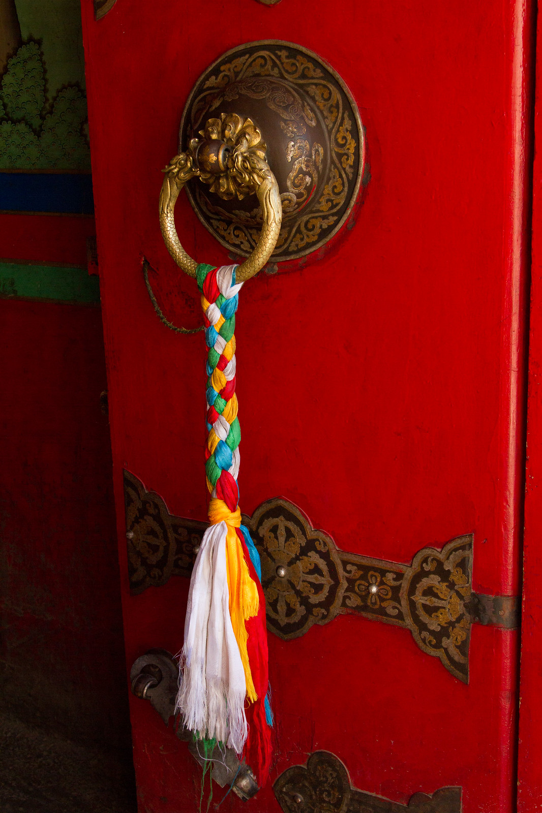 Ornate brass dragon door knocker on a red Tibetan temple door, hung with a braided rainbow-colored silk scarf.