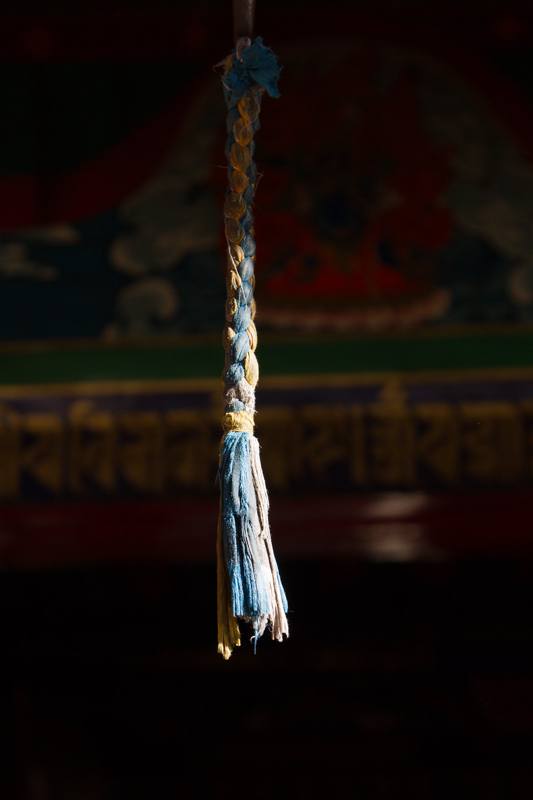 Braided blue and gold silk tassel hanging before a painted Tibetan monastery mural with gilded script.