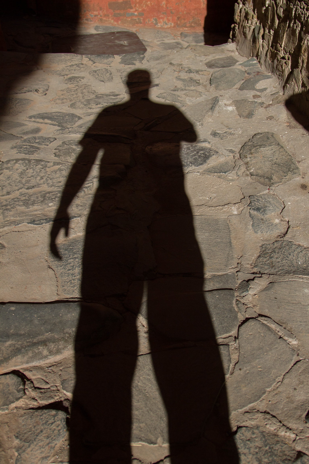 A silhouette shadow of a person standing, cast across a cracked flagstone courtyard beside a red wall.