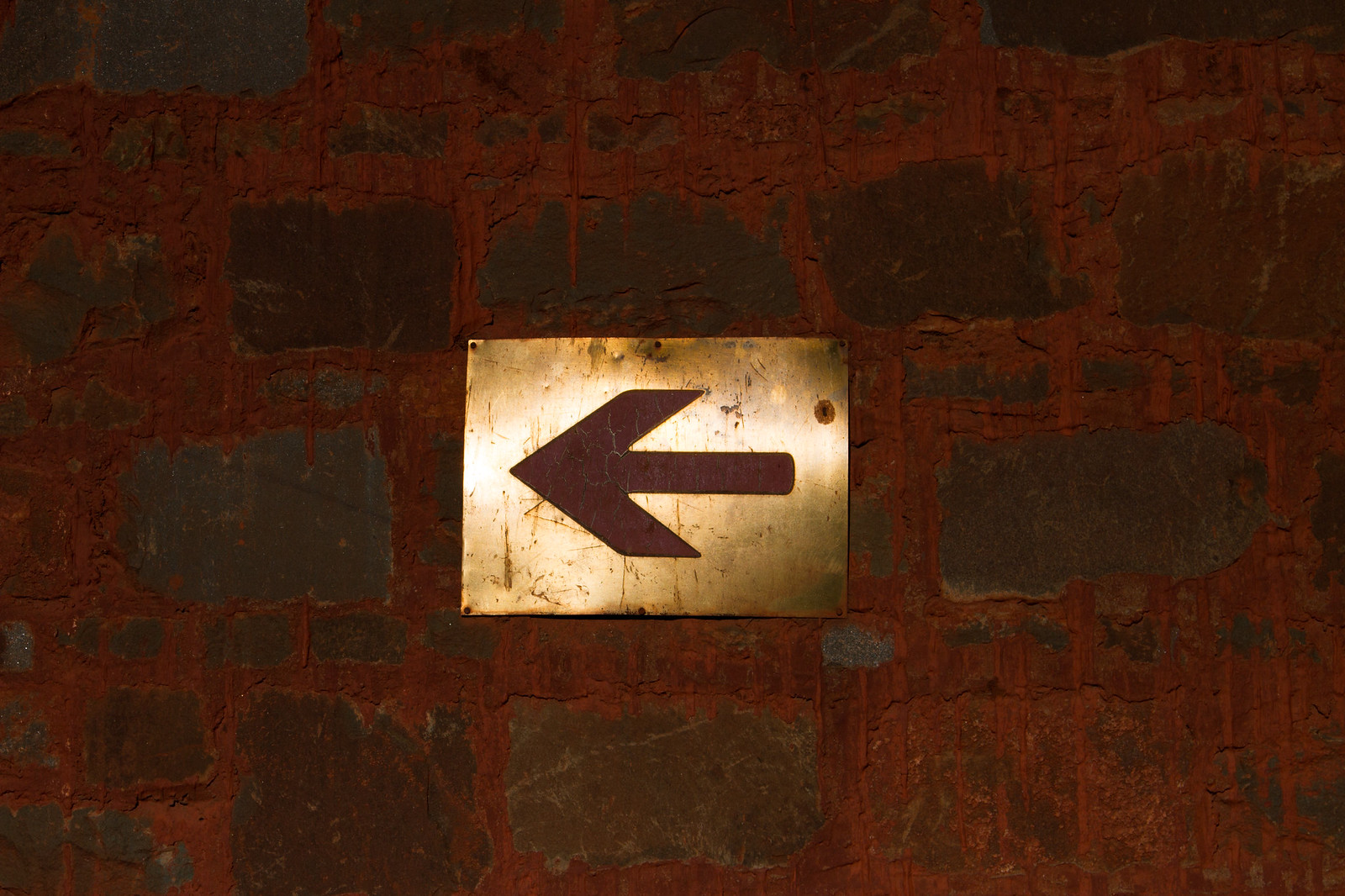 A tarnished brass plaque with a dark red left-pointing arrow, mounted on a deep red brick wall.