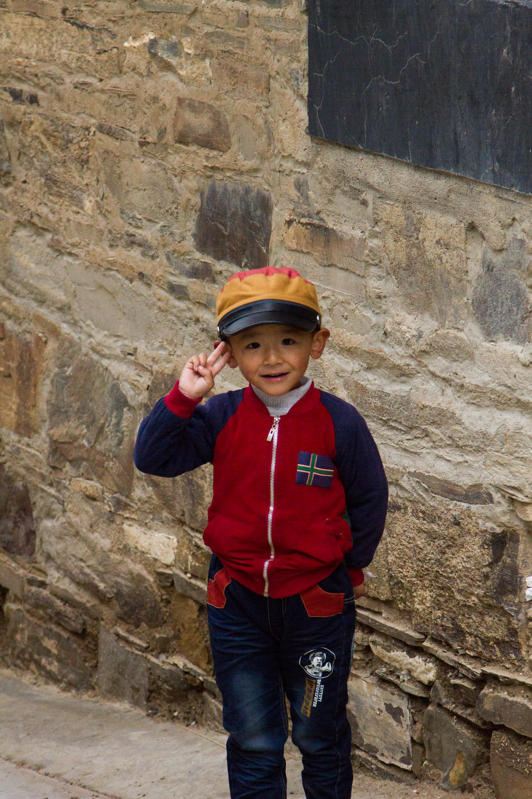 Smiling young boy in a yellow cap and red jacket poses against a stone wall.
