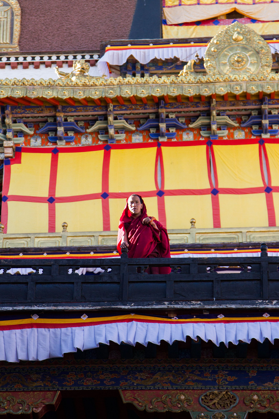A monk in crimson robes stands on an ornate gilded temple balcony draped with yellow and red banners.