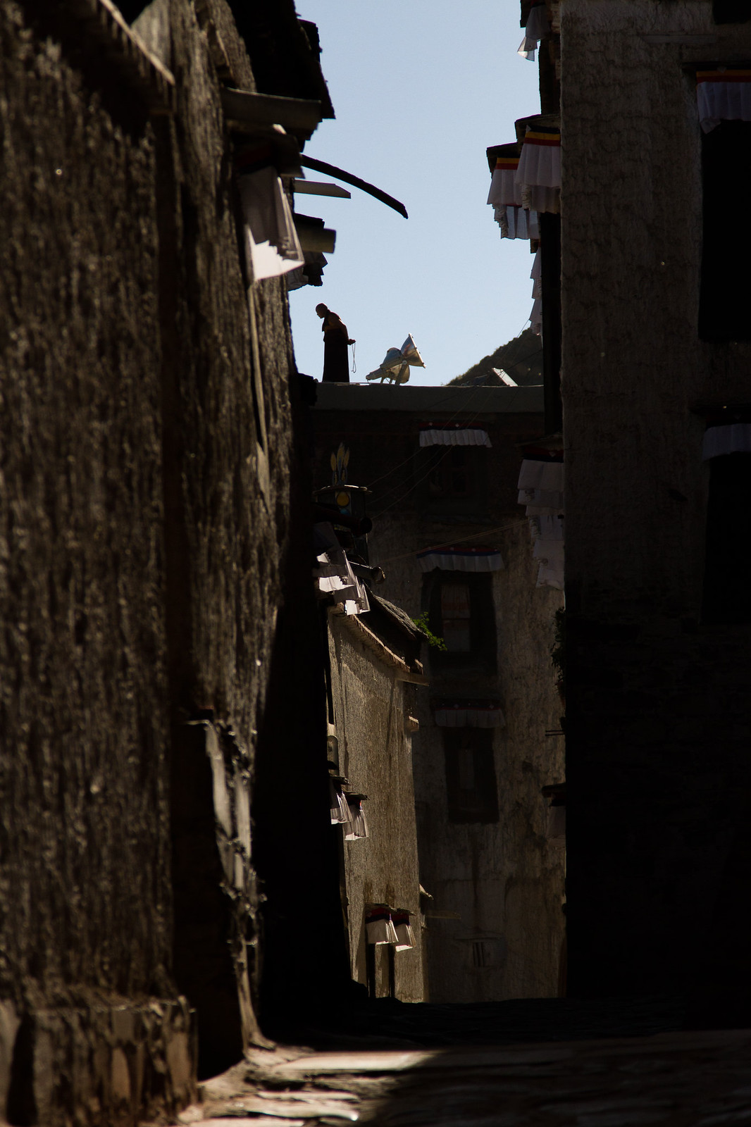 A robed monk stands on a sunlit rooftop above a shadowed Tibetan alley strung with white prayer cloths.