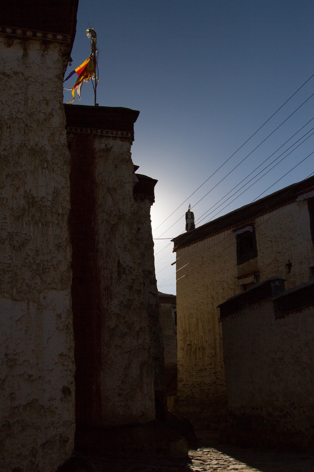Silhouetted whitewashed Tibetan buildings and a tattered prayer flag against the backlit sky at dusk.