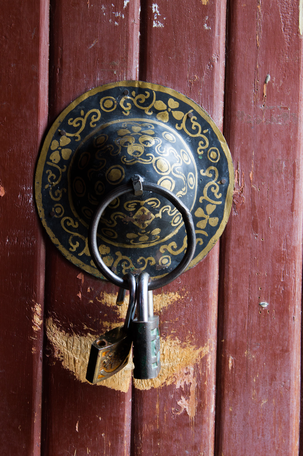 Ornate gold-and-black Tibetan door knocker with a ring holding two padlocks on a weathered red door.