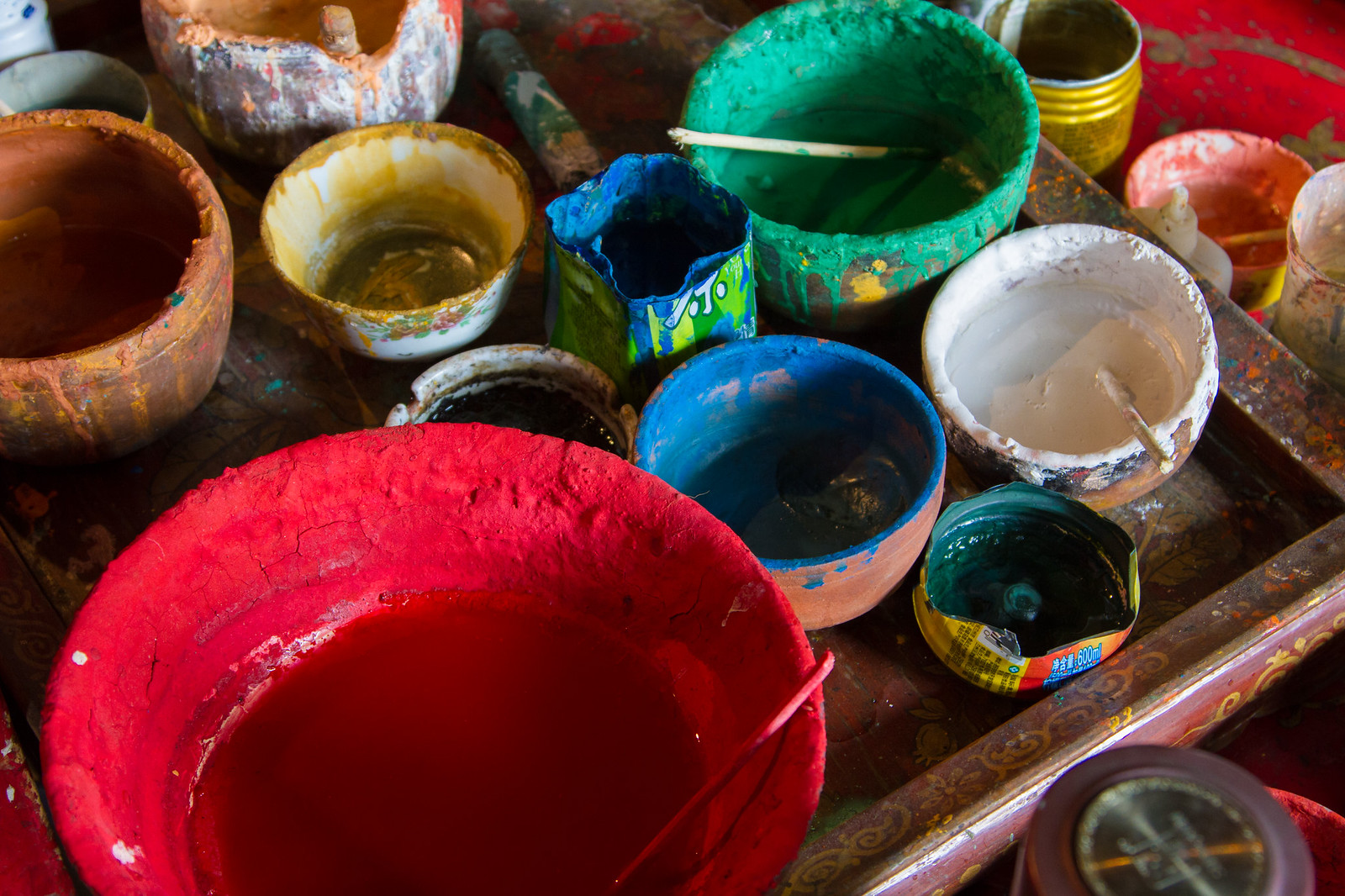 Colorful clay bowls of red, blue, green and yellow paint pigments on a worn Tibetan artist's tray.
