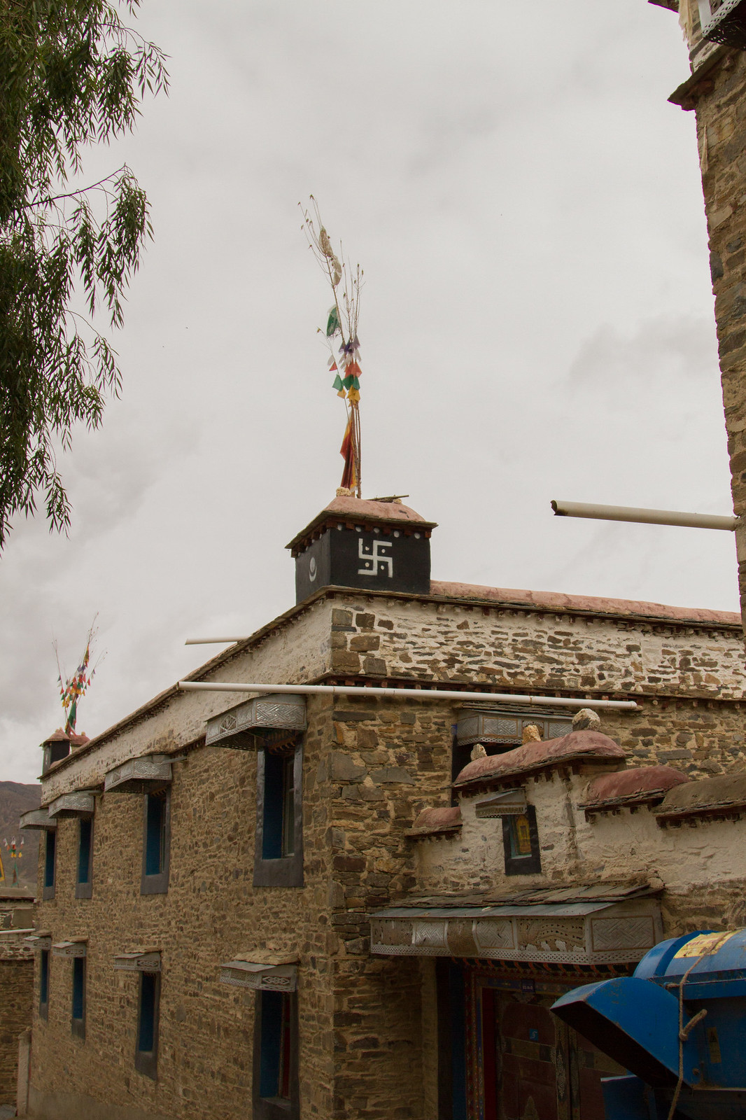 Traditional Tibetan stone house with a rooftop prayer-flag pole and a white Buddhist swastika symbol under a grey sky.