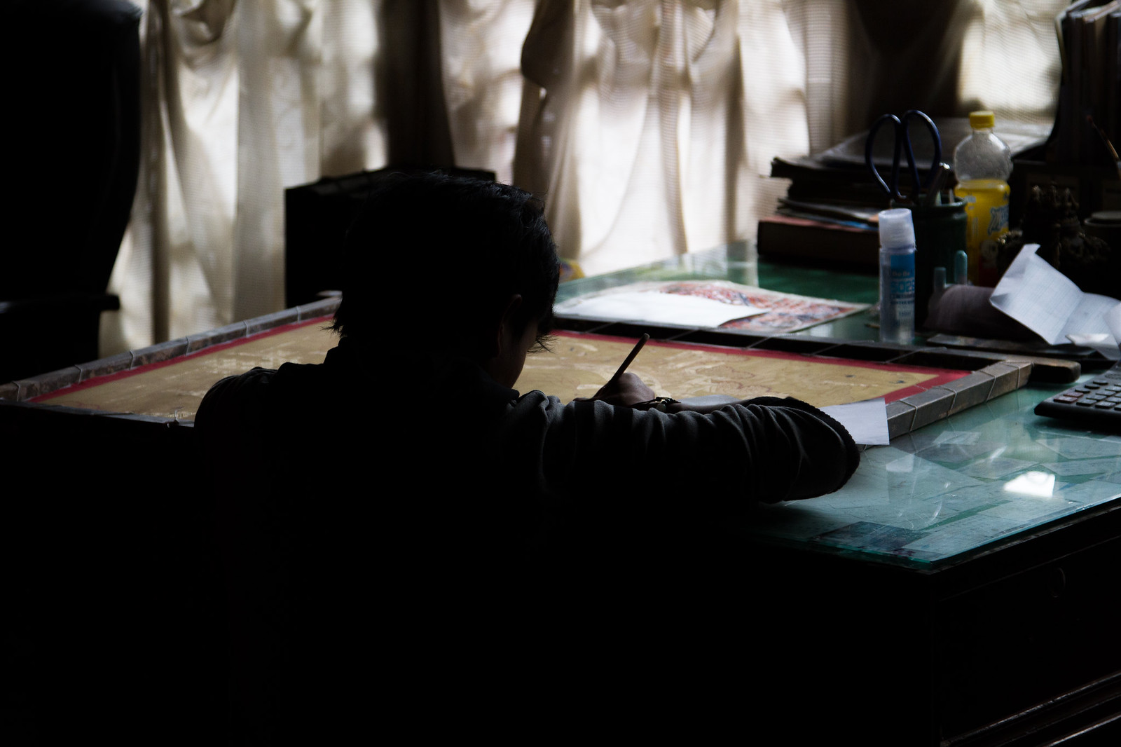 A silhouetted person paints a Thangka at a desk against sunlit white curtains in a Tibetan workshop.