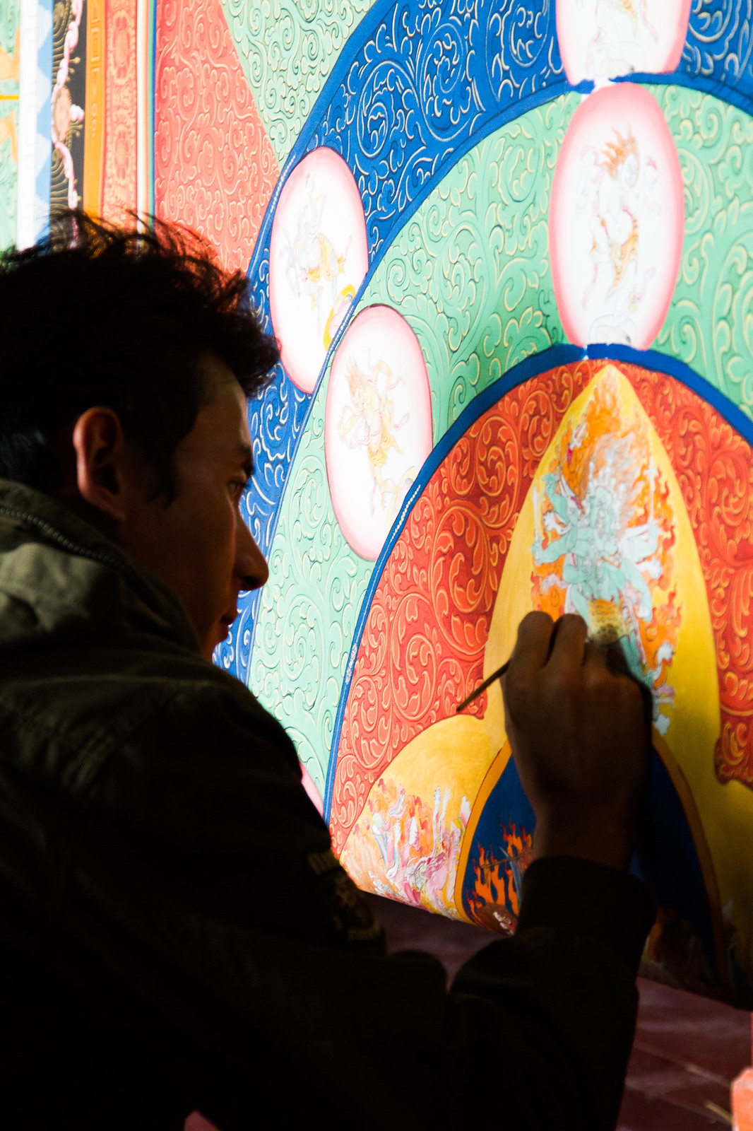 A young man in profile paints an intricate, colorful Tibetan Buddhist mandala thangka with a fine brush.