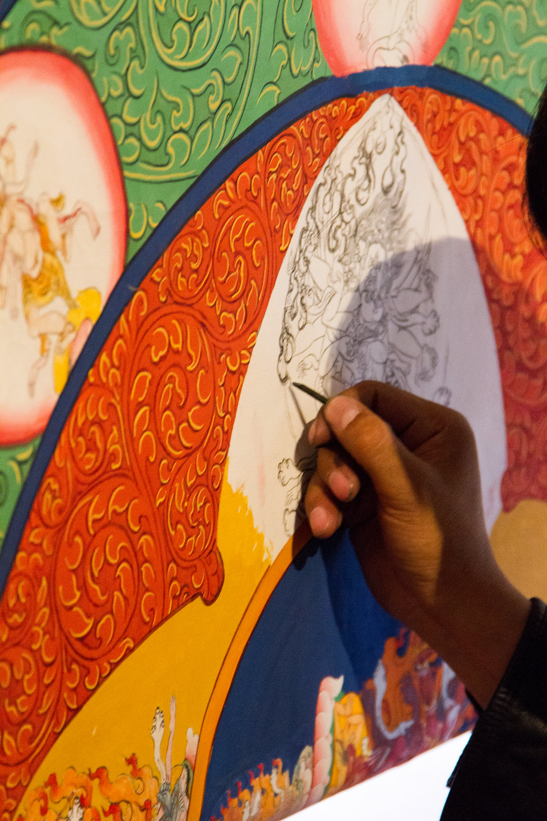 A hand uses a fine brush to ink intricate line-drawn deities on a vividly painted Tibetan thangka.